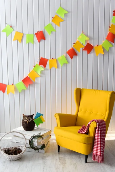 Interior of colorful unisex room for child. A neat white room with a bright yellow contrasting armchair, hand made toy owl and a wreath next to it, colourful flags garland hanging on the wall