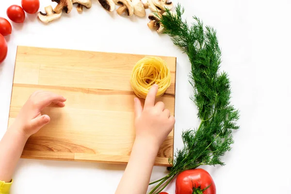 Child\'s hands touching the pasta nest on a wooden desk, vegetables and mushrooms around it. Cutting board, vegetables and Baby\'s hands. Top view, isolated on white background