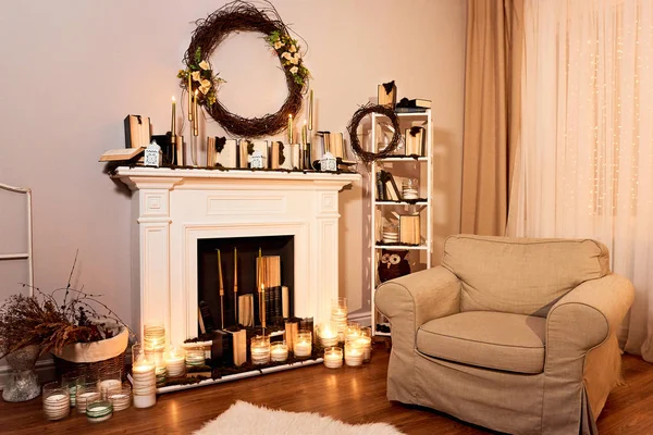 Modern designed spacious living room with autumn decorations. Fireplace with candles, cozy armchair, autumn wreath on the wall. Night windows at the background