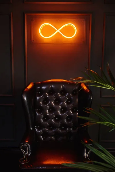 Leather armchair and a palm tree. Neon glowing sign with infinity and green wall.
