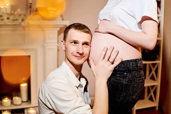 夫は妊娠中の妻を抱きしめる 家の中で素敵な妊娠中の妻の陽気な男抱擁腹の作物の写真と彼らの赤ちゃんを聞こうとします 脇を見ると — ストック写真