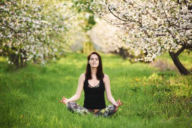 Kapalı gözlü Yogi kızı bahar bahçesinde yeşil çimlerde oturmuş Ardha Padmasana egzersizi yapıyor..