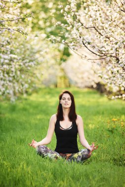 Kapalı gözlü Yogi kızı bahar bahçesinde yeşil çimlerde oturmuş Ardha Padmasana egzersizi yapıyor, tam boy. Dikey fotoğrafçılık