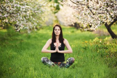 Çaylak gülümseyen, gözleri kapalı, spor giyimli, baharda yeşil çimlerde oturan, Lotus pozu veren, namaste ile yoga yapan bir kız..