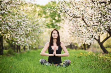 Uyum kavramı. Gülümseyen Yogi kız, gözleri kapalı spor kıyafetleri giyiyor, baharda yeşil çimlerde oturuyor çiçek açan bahçede, Lotus pozuyla yoga yapıyor, namaste ile tam boy..