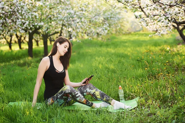 美丽迷人的女孩坐在盛开的花园里的绿色草坪上 一只手拿着手机 在无线耳机上听着音乐 面带微笑 放松冷静的概念 — 图库照片