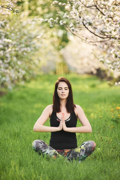Kapalı gözlü Yogi kızı bahar bahçesinde yeşil çimlerde oturuyor, yoga yapıyor, namaste ile egzersiz yapıyor, tam boy. Dikey fotoğrafçılık