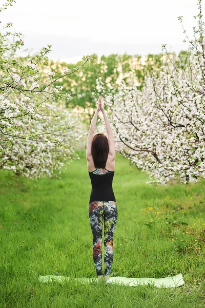 户外瑜伽练习 年轻的女人站在绿色的草坪上 在春花怒放的花园里 后视镜 手举起来在Namaste Mudra 平静和平静的心情 纵向摄影 — 图库照片