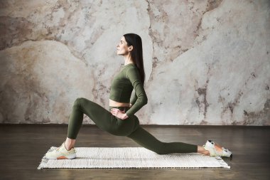 Uzun siyah saçlı, spor giyimli genç ve gülümseyen bir kadın, yoga stüdyosunda çalışıyor, duvar kenarındaki bej desenli arka planda çalışıyor, yoga egzersizi yapıyor, tam boy, yan görüş, spor yapıyor.