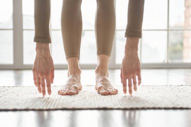Kadın büyük pencereye karşı açık renkli minder üzerinde duruyor, baş-diz egzersizi yapıyor, elleri ayakla kapatıyor, görünür damarlar ve kılcal damarlar, ev kavramında yoga yapan kadın öne eğik poz veriyor..
