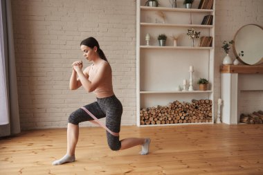 Bacak kasları için hamleler yapan genç bir kadın. Bacak halkaları için ilmek bantları kullanıyor. Kayış lastiğiyle yan yana egzersizler yapıyor. Kalça selülitleri için kalça kası aktivasyonu, tam boy.