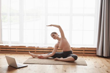 Spor giyimli genç bir kadın yoga yan bükme egzersizi yapıyor, evdeki bir laptoptan online eğitim videosu izleyerek pratik yapıyor. Sağlıklı yaşam tarzı, yoga, çevrimiçi öğrenme konsepti, boş alan
