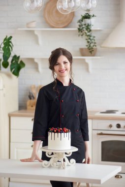 Gülümseyen ve kameraya bakan genç kadın şekerlemecinin portresi mutfakta tek katmanlı Drip Cake ile süslenmiş beyaz çikolata ganache ahududu, çilek, böğürtlen.
