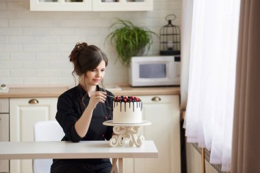 Siyahlı genç kadın pasta şefi masada oturmuş tek katmanlı Drip Cake 'i ahududulu, böğürtlenli ve yaban mersinli beyaz çikolatalı ganache ile süslüyor. Boşluğu Kopyala.