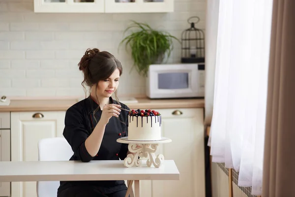 身穿黑衣服的年轻女糕点厨师坐在餐桌旁 在厨房里装饰着一层一层的滴滴蛋糕 上面涂满了覆盆子 黑莓和蓝莓 复制空间 — 图库照片