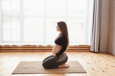 Genç hamile bir kadının dizlerinin üzerinde doğal bir halı üzerine oturup kalçasını esnetirken, karnını tutarken yoga egzersizinden sonra büyük pencereye bakarken yan görüntüsü. Yatay, tam uzunluk, kopyalama alanı.