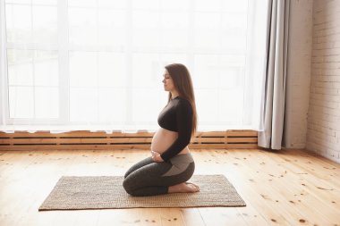 Genç hamile bir kadının dizlerinin üstüne çöküp kalçasını esnetme egzersizleri yapması, aşağıya bakması, yoga egzersizinden sonra dinlenmesi. Yatay, tam boy, kopyalama alanı, Hamilelik Yogası ve Fitness, Annelik