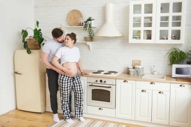 Ev kıyafetli müstakbel ebeveynler mutfakta durup birbirlerine bakıp öpüşüyorlar. Sağlıklı yaşam tarzı, ebeveynlik, annelik, annelik, çocuk beklentisi. Uzayı kopyala Tam uzunluk