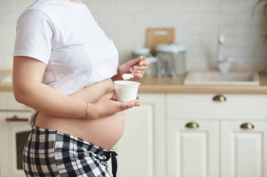 Ev kıyafetli, kimliği belirsiz, yakın plan sağlıklı genç bir kadın: beyaz tişört ve kareli pantolon, şirin göbekli, kahvaltıda beyaz Yunan yoğurdu tutan. Evde refah ve hamilelik kavramı.