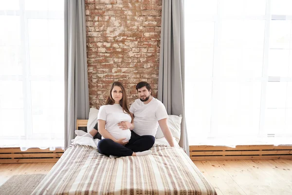 照片上 积极的丈夫和怀孕的妻子坐在白色的床上 看着相机 怀孕福利概念 在婚姻关系中复制空间 爱和支持 — 图库照片