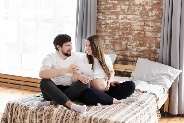 快乐的年轻孕妇和她的丈夫抱着超声波图像坐在床上在家 作为母亲和父母 家庭观念中的幸福 怀孕通知 特殊的家庭时刻 — 图库照片