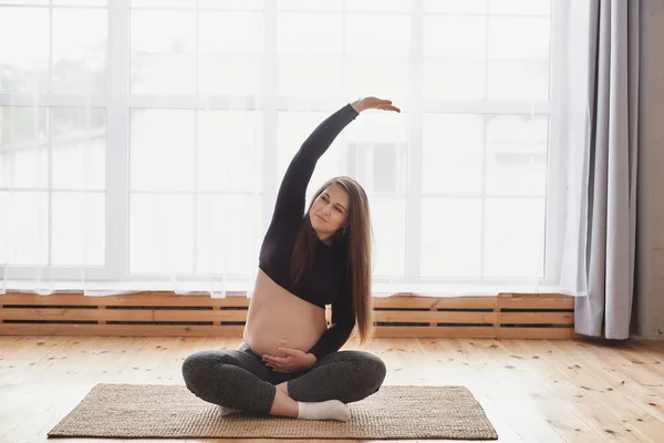 Portrait of smiling young pregnant woman in sports outfit sitting with crossed legs, she holding her tummy and bending aside with raised arm while practicing yoga at home. Motherhood, pregnancy, yoga