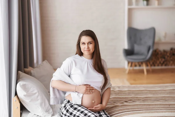 幸せな妊婦の家庭服のカメラを見て 白いTシャツと小切手パンツのベッドの上に座って 彼女のおなか 将来の母親の夢を抱擁 出生前 赤ちゃんの期待の概念 スペースのコピー — ストック写真