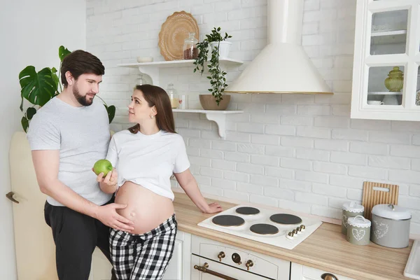 未来的父母穿着家居服站在厨房里拥抱 怀孕的妻子给丈夫一个绿色的苹果 生活方式 父母身份 对孩子的期望 复制空间 — 图库照片