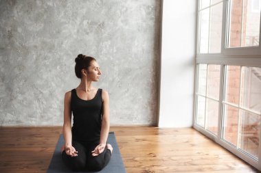 Orta yaşlı bir kadın yoga yapıyor, Sukhasana egzersizi yapıyor, rahat koltuk pozu veriyor, siyah spor kıyafetleri giyiyor ya da büyük pencere arkası olan bir yoga stüdyosu. Farkındalık ve sağlıklı yaşam tarzı kavramı.