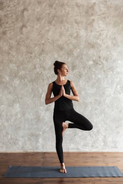 Yoga yapan güçlü, orta yaşlı bir kadın, başka yöne bakıyor, Vrksasana egzersizi yapıyor, ağaç pozisyonu yapıyor, spor yapıyor, yoga stüdyosunda gri duvarlı siyah spor giyim kuşamı giyiyor. Sağlıklı yaşam konsepti.