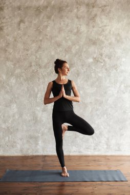 Yogi orta yaşlı, gülümseyen bir kadın yoga yapıyor, dikkatini başka tarafa veriyor, Vrksasana egzersizi yapıyor, ağaç duruşu yapıyor, spor yapıyor, evde gri duvarlı siyah spor giysiler giyiyor. Sağlıklı yaşam konsepti.