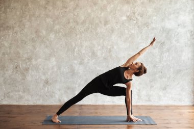 Orta yaşlı bir kadın yoga yapıyor, Utthita parsvakonasana egzersizi yapıyor, Genişletilmiş kenar açılı poz veriyor, yukarı bakıyor, spor yapıyor, siyah spor giyiniyor, kapalı alanda uzun, siyah pantolon giyiyor.