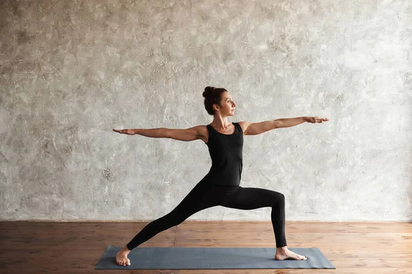 Orta yaşlı bir kadın yoga yapıyor, Virabhadrasana 2 egzersizi yapıyor, Warrior 2 pozu veriyor, spor yapıyor, siyah spor giyiniyor, kapalı alanda, yoga stüdyosunda gri duvar, kopyalama alanı, spor yapıyor.