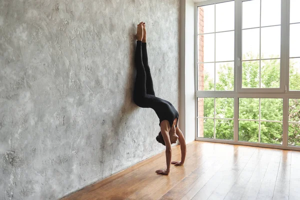 Woman practicing yoga, standing in Downward facing Tree Pose, in sportswear, doing Adho Mukha Vrksasana exercise near wall, at home or at yoga studio with big window, copy space, yoga studio, indoor