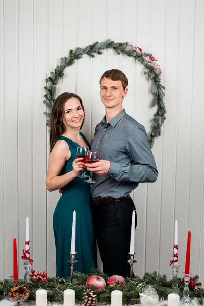 A family portrait of a hugging couple smiling for the camera clinking glasses. Beautiful couple in a decorated festive interior with a Christmas tree drinking wine. Looks at the camera and smiling.