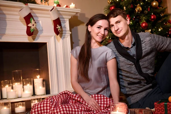 Portrait of gentle couple sitting near fireplace at home, romantic celebration of Christmas holidays, love. Newlyweds sitting closely to each other smiling happily, christmas atmosphere in the room.