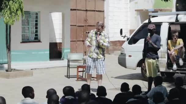 KISUMU, KENIA - 15 DE MAYO DE 2018: Joven africano y caucásico de pie, sosteniendo micrófonos y hablando con niños africanos — Vídeos de Stock