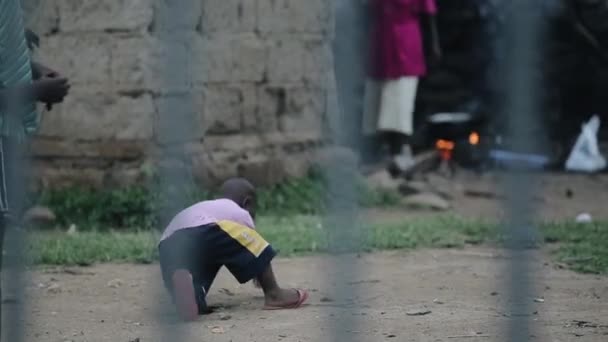 KISUMU,KENYA - MAY 21, 2018: View through the fence. Little african boy drawing with stick on the ground, play on backyard. — Stock Video