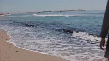 Sandy ile köpüklü dalgalar deniz kıyısı. Geri bir güzel görünümünü kapatmak yalınayak kumlu plaj boyunca yürüyüş kısa elbiseli kız