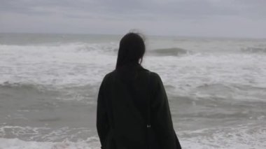 Geri denize bakarak koyu yeşil paltolu güzel bir kız görünümünü kapatın. Sonunda, o etrafında döner ve gülümsüyor. Rüzgarda çırpınan saç