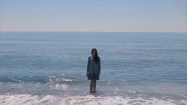 En vacker flicka i en våt kort klänning går ur vågiga havet och går mot kameran — Stockvideo
