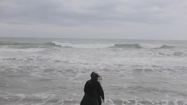 Een mooi meisje lopen vrij naar de golvende zee op een koude dag, raken het water, en loopt terug — Stockvideo