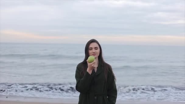 一个美丽的女孩在深绿色的大衣与绿色苹果在海边 — 图库视频影像