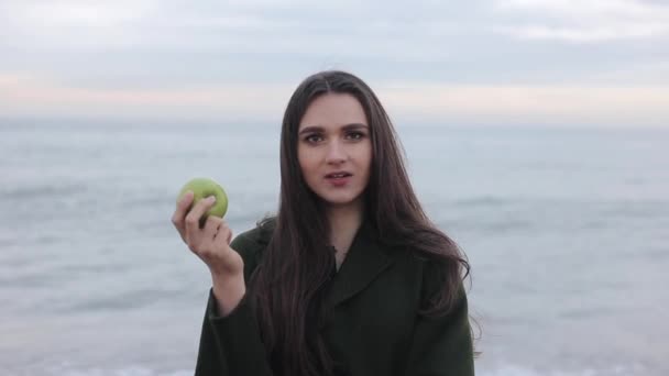 Güzel seksi kız sahilde taze elma yeme yakın bir bakış. Deniz kenarında yeşil paltolu bir kız — Stok video