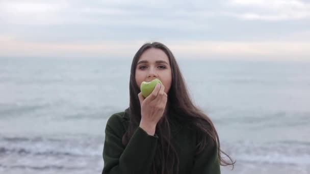 Um close-up de uma menina bonita em um casaco verde escuro mordendo uma maçã saborosa enquanto estava na praia no início da manhã — Vídeo de Stock