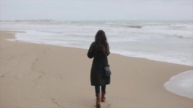 Sıcak giysiler giymiş genç bir esmer kumsalda yürür ve deniz görünümü bir bahar gününde sahiptir