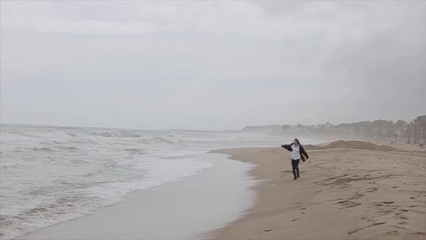 Uma menina bonita em roupas quentes corre ao longo da costa. Vista do mar com ondas espumosas em um dia ventoso — Vídeo de Stock