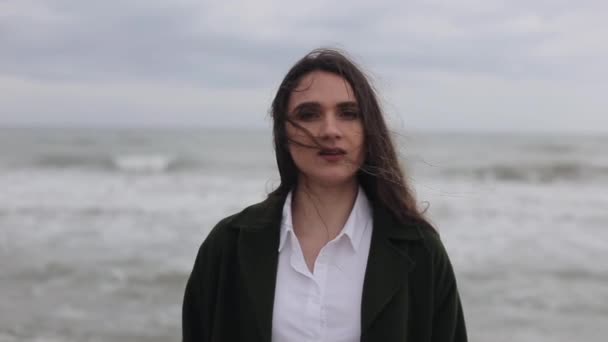 Een nauwe uitzicht op een mooi hijgend meisje gekleed in een wit overhemd en een donker groene jas permanent op het strand met de zee op de achtergrond — Stockvideo