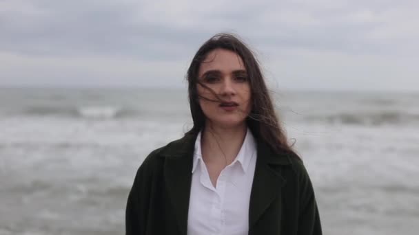 Eine Nahaufnahme eines schönen hechelnden Mädchens in weißem Hemd und dunkelgrünem Mantel, das am Strand steht, mit dem Meer im Hintergrund — Stockvideo