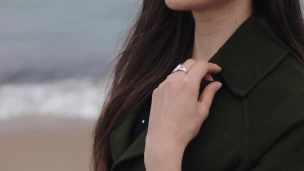 A close-up of a beautiful girl covering her neck with a dark green coat and stroking her long dark hair. Blurred background of the sea and foamy waves — Stock Video